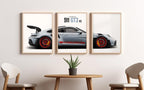 A three-panel framed wall art print set showing a silver Porsche 911 GT3 RS, displayed in a modern room setting above a wooden table with two chairs.