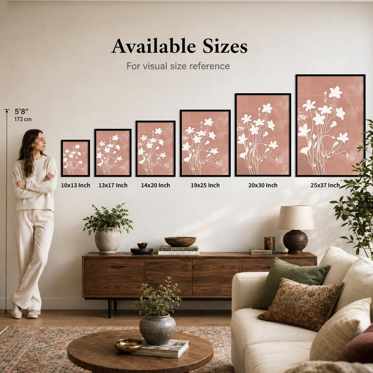 A lifestyle room scene showing a woman standing beside a row of framed floral wall art prints with text reading 'Available Sizes' and size labels from 10x13 inch to 25x37 inch. The room includes a sofa, console table, lamp, plants, and rug.